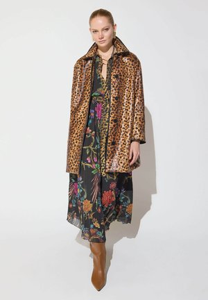 Mujer con abrigo de estampado de leopardo sobre un vestido floral negro con botas marrones de tacón, de pie frente a un fondo blanco liso.