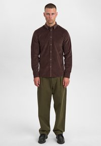 Brun kordfløjsbutton-up skjorte med lange ærmer, parret med løse olivengrønne bukser og sorte sko. Simpelt, struktureret stofdesign.