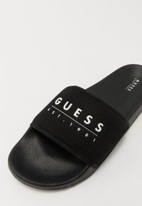 Fekete csúszka szandál texturált pánttal, amelyen fehér "GUESS EST. 1981" logó található. Sima fekete talp, minimalista dizájn és rugalmas anyag jellemzi.