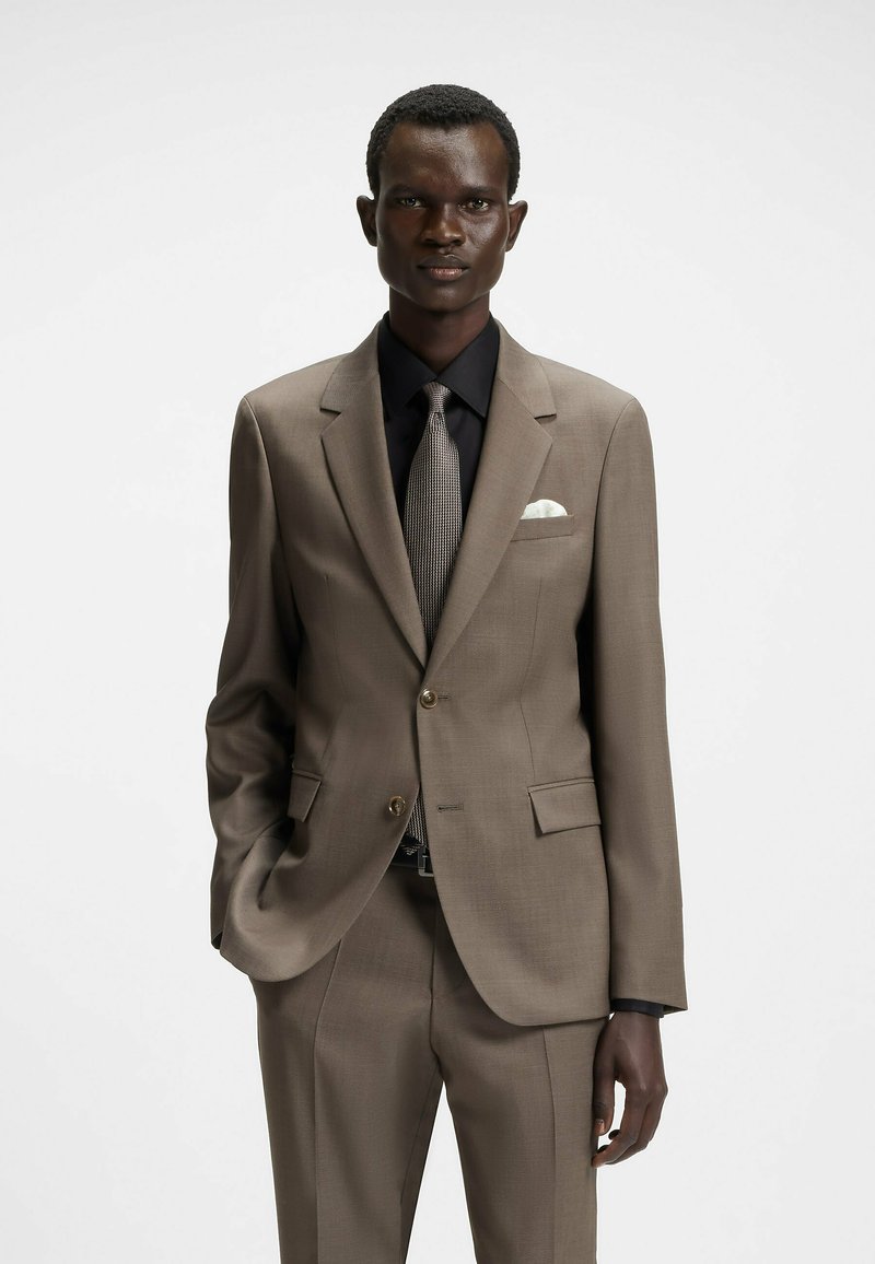 Brąski dopasowany garnitur z klapami w kształcie notched, z jednym guzikiem i dwiema przednimi kieszeniami. Połączony z czarną koszulą i teksturowanym szarym krawatem. Biała chusteczka w kieszeni.