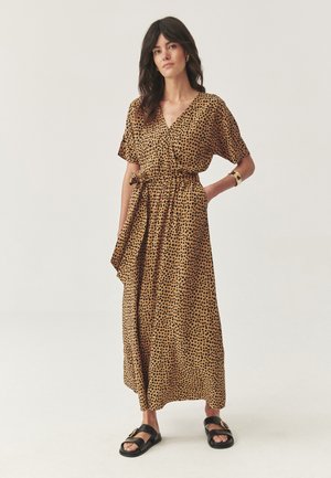 Femme debout portant une robe enveloppante beige à pois noirs, à manches courtes, avec une ceinture à nouer, un bracelet en or et des sandales noires à boucles.
