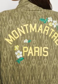 Olijfgroen jack met geborduurde tekst "MONTMARTRE PARIS" in het geel, met bloemaccenten van witte en gele bloemen. Gegolfd patroon door het hele jack.