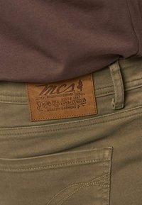 Καφέ δερμάτινο πλακίδιο με το κείμενο "MCA Rugged and Refined", ραμμένο στην πίσω πλευρά της ελιάς παντελονιών κάτω από ένα καφέ πουκάμισο.