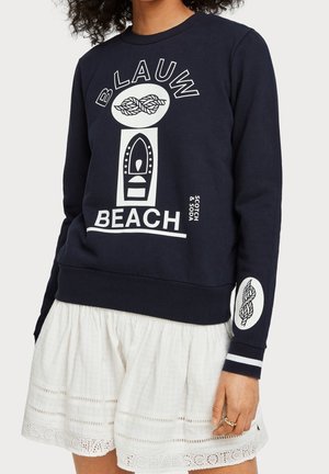 Kobieta w granatowej bluzie z białym graficznym napisem „BLAUW BEACH” oraz wzorami sznura i łodzi, zestawiona z białą spódnicą.