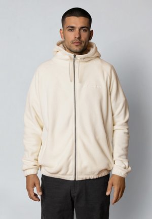 Mand med kort mørkt hår iført en cremefarvet fleece-hoodie med lynlås og sorte bukser, stående foran en ensartet lys baggrund.