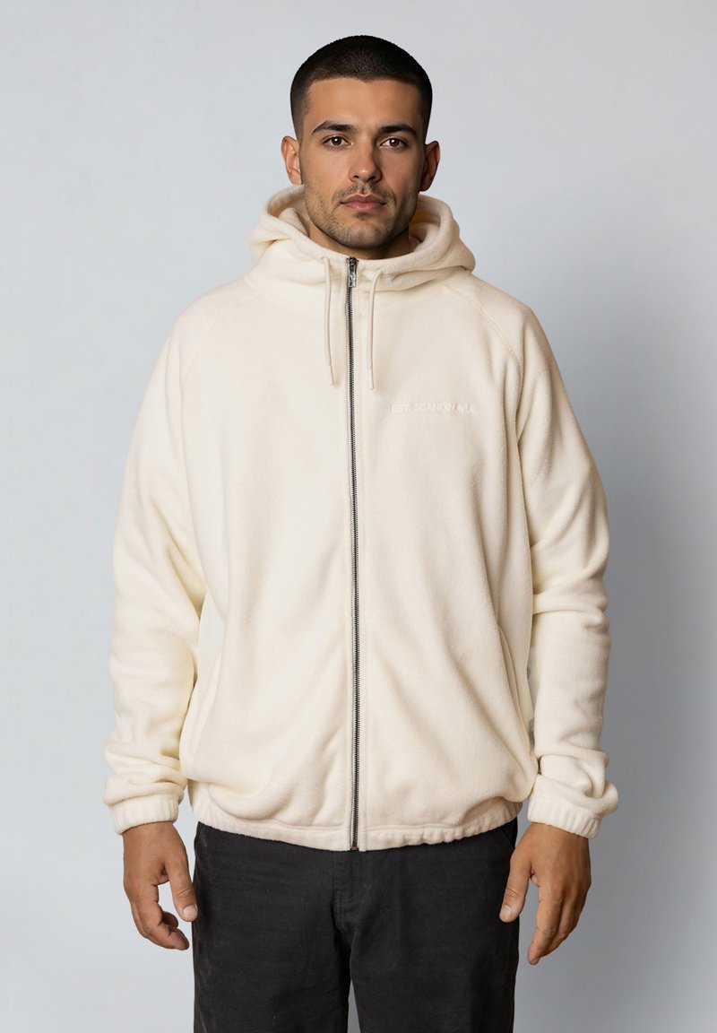 Mężczyzna o krótkich ciemnych włosach, ubrany w kremową bluzę polarową na zamek i czarne spodnie, stojący na jasnym, jednolitym tle.