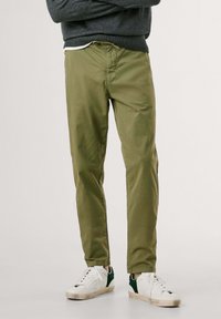 Chinos vert olive avec une coupe ajustée, fabriqués en tissu lisse. Associés à des baskets blanches avec des accents verts.