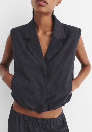 Kobieta nosząca bezrękawnik ciemnoszary żakiet z głębokim dekoltem w serek i elastycznym mankietem, w połączeniu z dopasowanymi ciemnoszarymi spodniami, ręce w kieszeniach.