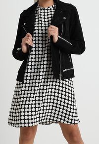 Svart mockabikerjacka med silverdragkedjor över en vit klänning med svarta prickar, korta ärmar och en utsvängd fåll.