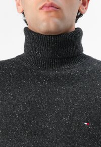 Temno siva turtlenecka pulover, izdelan iz teksturirane pletenine s pikčasto strukturo; ima majhen logotip v spodnjem kotičku.