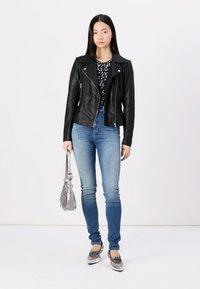 Chaqueta de motocicleta de cuero negro sobre una blusa de patrón en blanco y negro, combinada con jeans ajustados de color azul claro y zapatos plateados. Incluye un bolso gris.