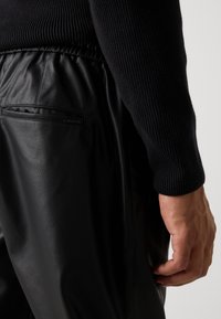 Primer plano de una persona que lleva pantalones de cuero sintético negro y una camiseta de manga larga negra con canalé, con un bolsillo trasero visible y una mano relajada.