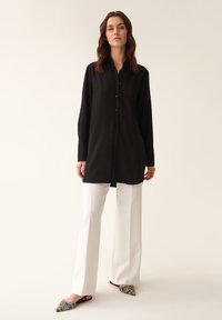 Schwarzes Langarmshirt mit Knopfleiste und lockerer Passform, kombiniert mit weißen, weit geschnittenen Hosen und schwarzen, gemusterten Slippers.