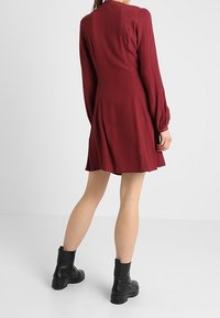 Burgundin pitkähihaine mekko, jossa on levenevä helma, valmistettu pehmeästä kankaasta, yhdistettynä mustiin nilkkasaappaisiin. Yksinkertainen muotoilu, jossa on minimalisti yksityiskohtia.