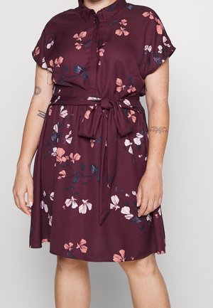Woman wearing a knee-length maroon dress with pink and white floral prints and a tied waist belt, showing arm tattoos and hands relaxed by sides.