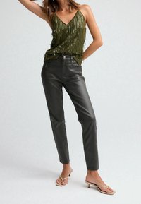 Top senza maniche color verde oliva con dettagli in frange metalliche, abbinato a pantaloni attillati in finta pelle grigia scura e sandali con tacco beige.