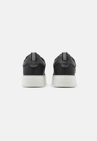 Adidași negri cu un superior din piele și plasă, având o talpă din cauciuc alb și un tab cu dungi la călcâi. Logo embossat pe călcâi.