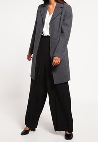 Cappotto di lana grigio con un design sartoriale, grande colletto e bottoni frontali, abbinato a pantaloni larghi neri e scarpe nere.