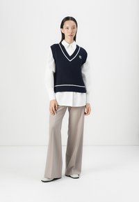 Navy ribbed knit vest with white trim, layered over a white button-up shirt. Model wears loose beige trousers and light-colored shoes.
