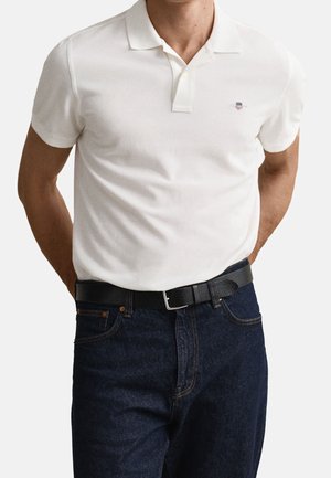Mand iført hvid kortærmet polo-shirt med lille logo på brystet, mørkeblå jeans og sort læderbælte med sølvspænde.