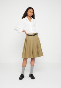 White button-up shirt with rolled sleeves, paired with a pleated khaki skirt, black belt, gray socks, and black loafers. Minimal background.