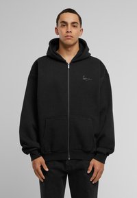 Black zip-up hoodie made of soft fabric with a kangaroo pocket, featuring a signature logo on the chest and a drawstring hood.