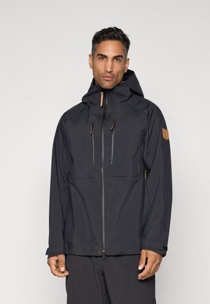 Man wearing black waterproof hooded jacket with front zippers and adjustable cuffs, standing against plain light background.