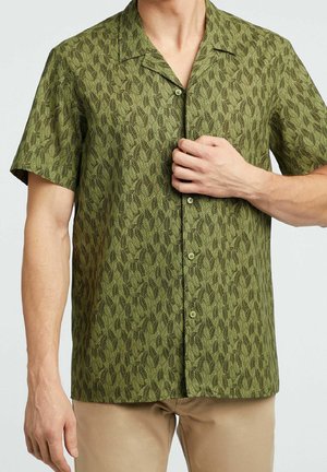 Mężczyzna nosi oliwkowo-zieloną koszulę z krótkim rękawem z ciemnym wzorem liści, w połączeniu z beżowymi spodniami, ręka poprawia przód koszuli.