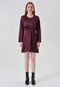 Abito in satin borgogna con maniche lunghe e vita arricciata. Presenta una parte anteriore drappeggiata, lunghezza sopra il ginocchio, abbinato a stivaletti neri con tacco.