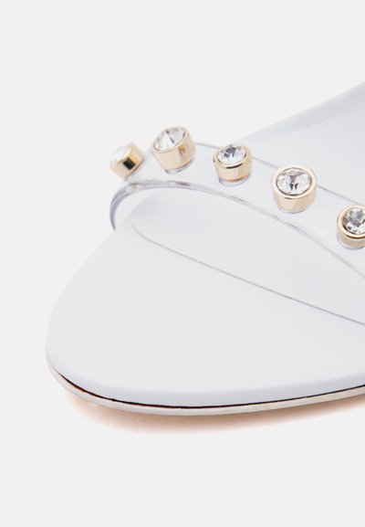 Λευκό παπούτσι με μυτερή μύτη και διάφανο λουράκι που διαθέτει χρυσά στοιχεία με στρογγυλά διάφανα πολύτιμα πετράδια.