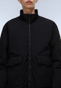 Giacca nera con zip e colletto alto, dotata di due tasche frontali e tessuto testurizzato. Dettaglio notevole include una toppa rotonda con logo sulla manica.