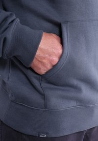 Sweat-shirt gris avec poignets et ourlet côtelés, doté d'une poche avant. Texture douce avec une étiquette de logo visible en bas.