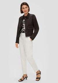 Zwarte jas met een knoopsluiting, twee voorzakken, gedragen over een zwart-wit graphic t-shirt, gecombineerd met witte broek en zwarte sandalen.