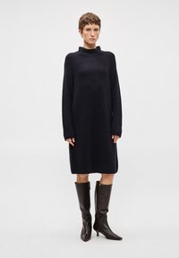 Πλεκτό μίντι φόρεμα σε σκούρο ναυτικό με ψηλό γιακά και μακριά μανίκια, συνδυασμένο με γυαλιστερές καφέ δερμάτινες μπότες μέχρι το γόνατο και μυτερά δάχτυλα.