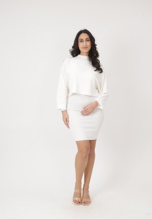 Kobieta o długich ciemnych włosach, ubrana w biały krótki sweter z długim rękawem i dopasowaną białą spódnicę, stojąca z jedną ręką na biodrze, neutralne tło.