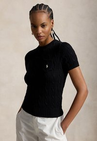 Mujer con cabello trenzado que lleva un suéter de punto trenzado negro de manga corta con un pequeño logo en el pecho y pantalones blancos, con las manos en los bolsillos.