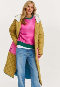 Prešívaný dlhý kabát v horčicovo žltej farbe s šedou kapucňou, vrstvený cez ružový pruhovaný sveter a modré džínsy. Obsahuje farebné látkové akcenty.