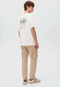 Ung mand iført en hvid grafisk T-shirt med løbesko, beige cargo-bukser og beige sneakers, stående sidelæns på en ensartet lys baggrund.