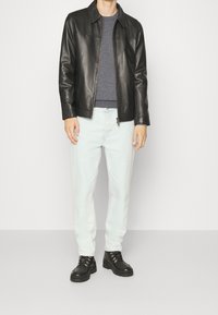 Schwarze Lederjacke mit Reißverschluss vorne und flachem Kragen, getragen über einem grauen Hemd. Hellblaue Jeans und schwarze Schnürstiefel runden das Outfit ab.