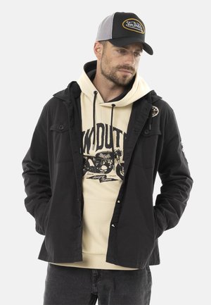 Homme portant une casquette Von Dutch noire et blanche, un sweat à capuche crème avec un motif de moto, une veste noire et un jean foncé, debout les mains dans les poches.