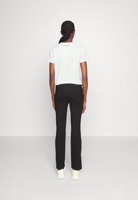 Vit kortärmad t-shirt och svarta tajta byxor, sedd bakifrån. T-shirten har en logotyp på den övre delen av ryggen. Bär vita sneakers.