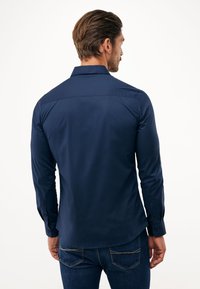 Camicia a manica lunga blu navy con design aderente, polsini con bottoni e texture liscia. Presenta un colletto classico e dettagli sullo sprone posteriore.