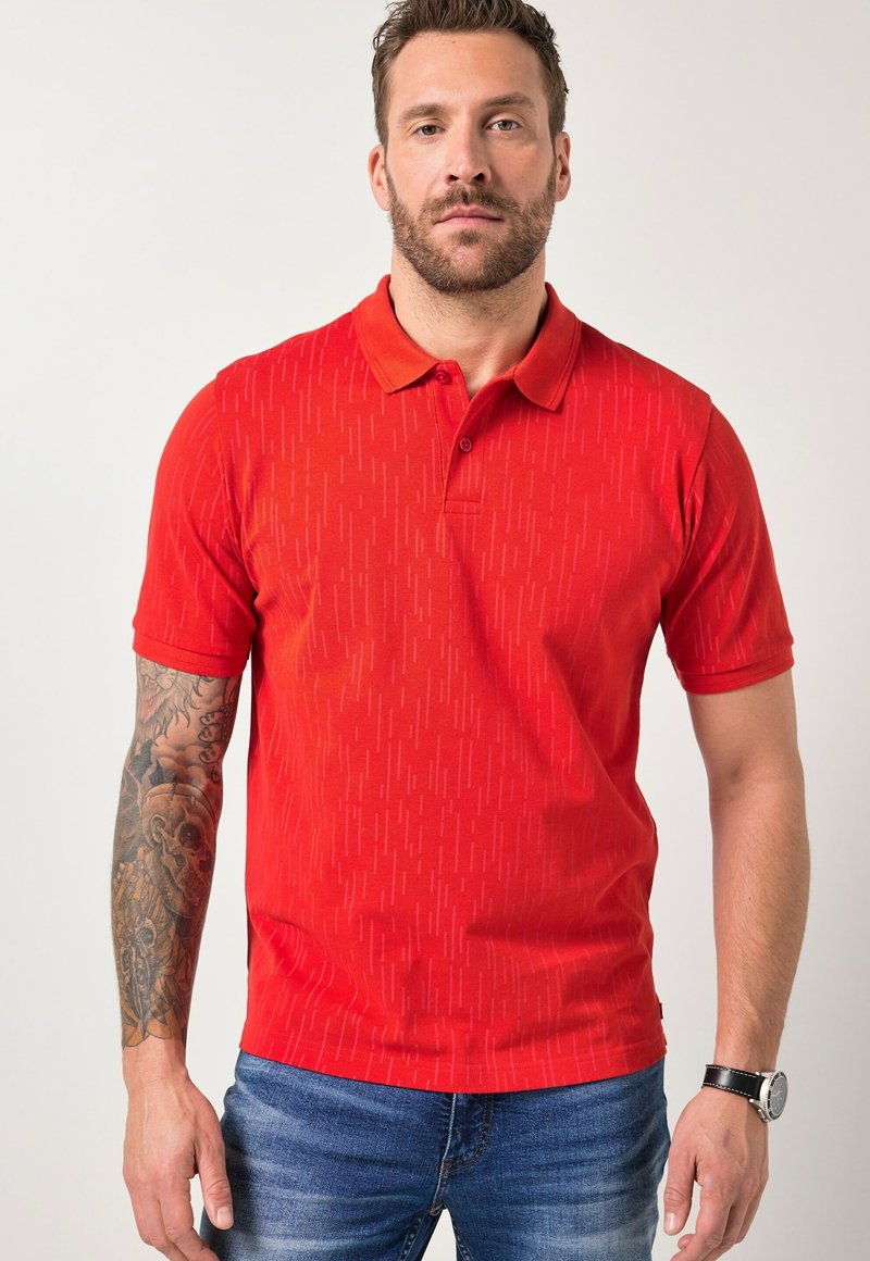 Mann mit Bart, der ein rot gemustertes Poloshirt, blaue Jeans, eine Uhr am linken Handgelenk und einen detaillierten Tattoo-Ärmel am rechten Arm trägt.