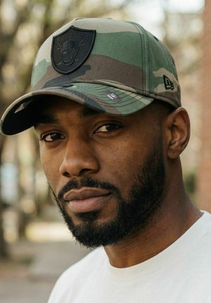 Mann mit Bart, der eine Camouflage-Raiders-Kappe und ein weißes Hemd trägt, blickt direkt in die Kamera vor einem unscharfen Außenhintergrund.