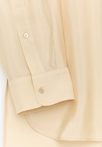 Chemise beige à manches longues avec une texture lisse, un poignet boutonné et un ourlet arrondi. Le tissu a un léger éclat et une finition douce.