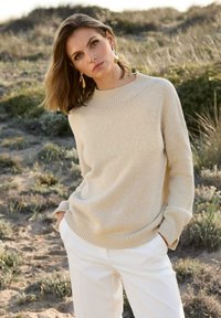 Beige Strickpullover mit Rundhalsausschnitt, überschnittenen Schultern und gerippten Bündchen. Kombiniert mit weißen Hosen vor einem natürlichen Hintergrund.