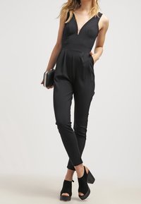 Svart jumpsuit med djup V-ringning, figurnära liv, avsmalnande ben och sidofickor. I kombination med svarta öppna plattformsskor.