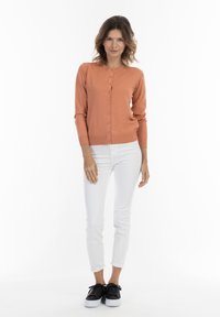 Cardigan pesca con scollo rotondo e chiusura a bottoni, abbinato a jeans skinny bianchi e sneakers nere, su uno sfondo bianco semplice.