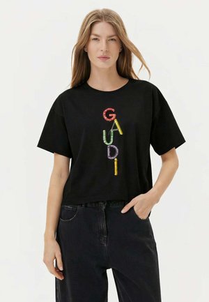 Donna con lunghi capelli biondi che indossa una maglietta nera con la parola "GAUDI" scritta in lettere colorate e perline, abbinata a pantaloni scuri.