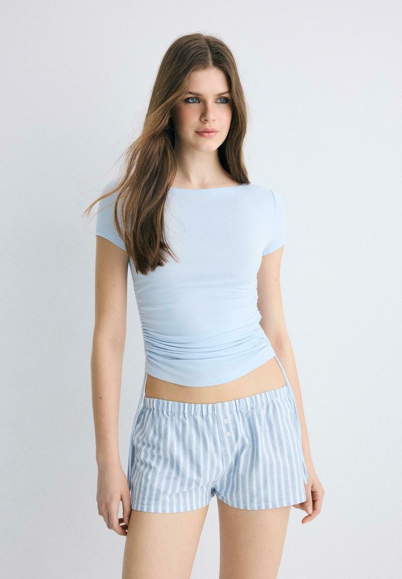 Jeune femme aux longs cheveux bruns portant un haut ajusté bleu clair et un short rayé bleu et blanc, sur un fond uni.
